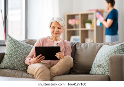 Technology, Old Age And People Concept - Happy Senior Woman With Tablet Pc Computer And Housekeeper Cleaning At Home