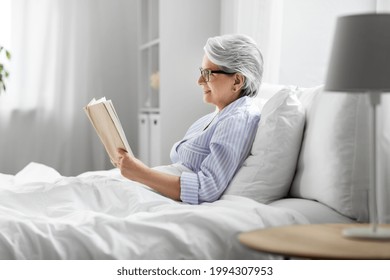 technology, old age and people concept - happy smiling senior woman in glasses reading book in bed at home bedroom - Powered by Shutterstock