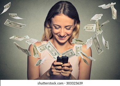 Technology online banking money transfer, e-commerce concept. Happy young woman using smartphone with dollar bills flying away from screen isolated on gray wall office background.