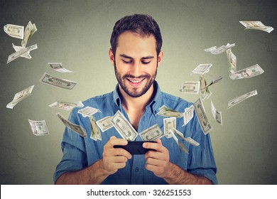 Technologie de transfert d'argent bancaire en ligne, concept de commerce électronique. Joyeux jeune homme utilisant un smartphone avec des billets de dollars s'envolant de l'écran isolé sur fond gris du bureau.