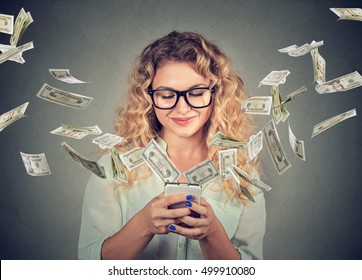 Technology online banking money transfer, e-commerce concept. Happy young woman using smartphone with dollar bills flying away from screen isolated on gray wall background
