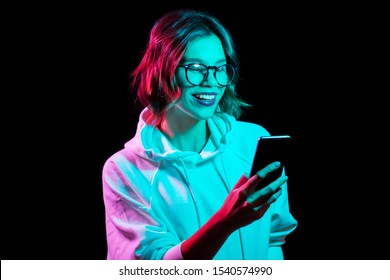 Technology And People Concept - Happy Young Woman Wearing Hoodie And Using Smartphone In Neon Lights Over Black Background