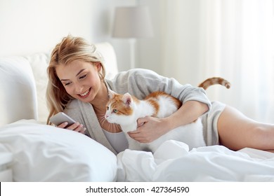 Technology, Pets, Communication And People Concept - Happy Young Woman With Cat And Smartphone Texting Message In Bed At Home