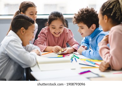 Technology And Pupils Concept. Group of excited multicultural happy junior children sitting at table and using smartphone, playing online mobile games. Modern Device, Gadget Addiction