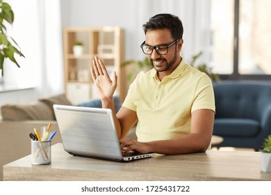 technology, remote job and lifestyle concept - happy indian man in glasses with laptop computer having video chat and waving hand at home office