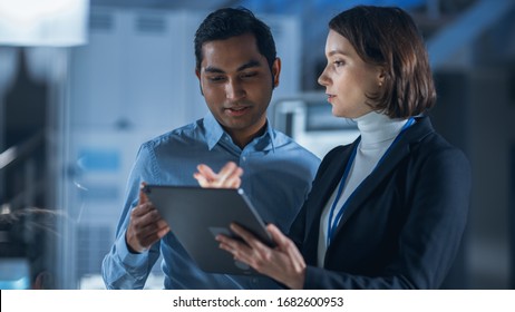 In Technology Research Facility: Female Project Manager Talks With Chief Engineer, They Consult Tablet Computer. Team Of Industrial Engineers, Developers Work On Engine Design Using Computers