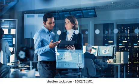 In Technology Research Facility: Female Project Manager Talks With Chief Engineer, They Consult Tablet Computer. Team Of Industrial Engineers, Developers Work On Engine Design Using Computers