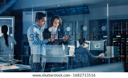 In Technology Research Facility: Female Project Manager Talks With Chief Engineer, they Consult Tablet Computer. Team of Industrial Engineers, Developers Work on Engine Design Using Computers