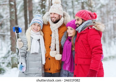 Technology, Season, Friendship And People Concept - Group Of Smiling Men And Women Taking Selfie With Smartphone And Monopod In Winter Forest