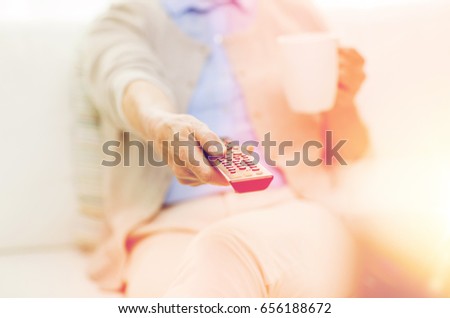 technology, television, age and people concept - close up of senior woman watching tv, drinking tea and changing channels by remote control at home