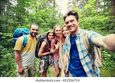 Technology, Travel, Tourism, Hike And People Concept - Group Of Smiling Friends Walking With Backpacks Taking Selfie By Smartphone Or Camera In Woods