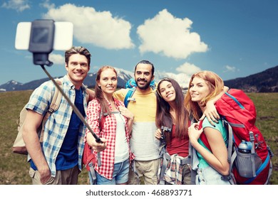 Technology, Travel, Tourism, Hike And People Concept - Group Of Smiling Friends Walking With Backpacks Taking Picture By Smartphone On Selfie Stick Over Natural Background