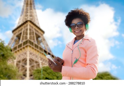 Technology, Travel, Tourism And People Concept - Smiling African American Young Woman Or Teenage Girl With Smartphone And Headphones Listening To Music Over Eiffel Tower Background