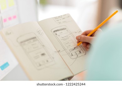 technology, user interface design and people concept - hand of ui designer or developer drawing smartphone sketches in notebook at office