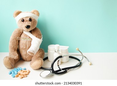 A teddy bear with a broken paw and a sore head sits on a turquoise background, next to pills, vitamins and a stotoscope.  The concept of children's medicine.  Banner place for copy text.