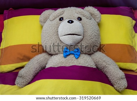 Teddy Bear

Close up of beautiful teddy bear lying in bed covered with blanket and ready to sleep.
He is wearing nice blue bow tie. Sheets looks like rainbow with  yellow, orange and purple colors.