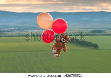 Teddy bear flying to the sky with a big yellow and red balloons.  The bear flying over the fields. 