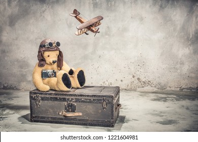 Teddy Bear With Leather Aviator's Hat And Goggles Sitting On Vintage Old Aged Classic Travel Trunk Circa 1900s With Film Camera And Flying Toy Plane. Travel By Air Concept. Retro Style Filtered Photo