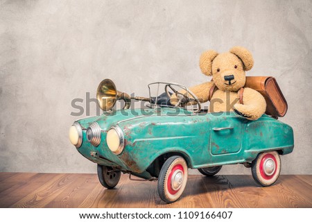 Teddy Bear and old leather school bag driving in rusty retro turquoise toy pedal car with classic brass klaxon in front concrete textured wall background. Vintage style filtered photo