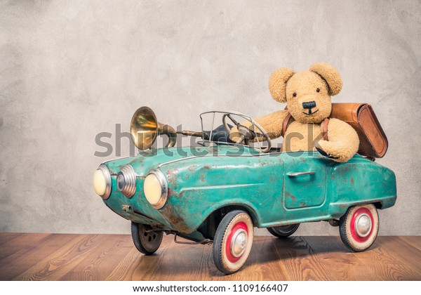 Teddy Bear and old leather school bag driving in rusty retro turquoise toy pedal car with classic brass klaxon in front concrete textured wall background. Vintage style filtered photo