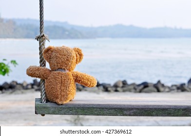 Teddy Bear sitting on the wooden swing with blue sea and sky background. Concept about loneliness.