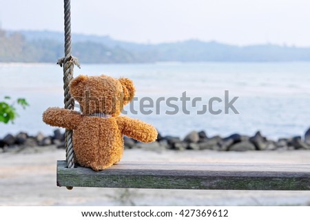 Teddy Bear sitting on the wooden swing with blue sea and sky background. Concept about loneliness.