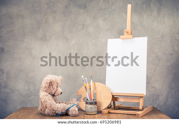 Teddy Bear toy, portable desk easel for painting\
with canvas blank, brushes and artist\'s palette on wooden table\
front concrete wall background. Retro instagram old style filtered\
photo
