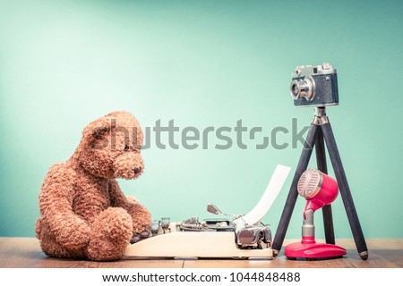 Teddy Bear toy sitting on old wooden desk with retro typewriter, outdated film photo camera on tripod and microphone front mint green wall background. Blogging concept. Vintage style filtered photo