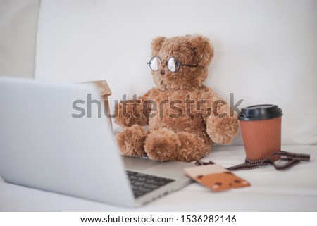 Teddy bear working concept. A cute teddy bear wearing glasses working in bed with a grey laptop and a cup of hot coffee