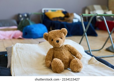 Teddybear of brown color sitting on bed done with white cotton blanket of refugee child in large room prepared for migrants