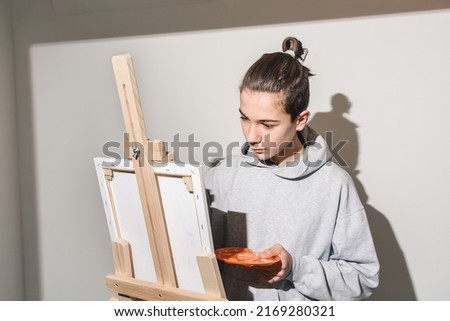 Teen artist intently paints picture on easel and holds brush. Child holds an orange palette in studio. Direct view, harsh light and uniform white wall. Guy in casual clothes. Copy Space, Soft focus