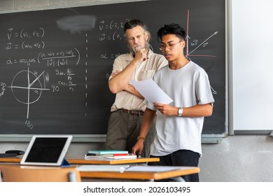 Teen Asian Boy High School Student Giving A Presentation In Class While Caucasian Mature Adult Teacher Observes His Notes. Education Concept.