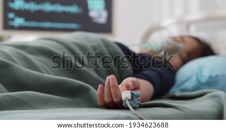 Teen asian girl in oxygen mask and oximeter sleeping in hospital bed. Portrait of unconscious child lying in bed and suffering from coronavirus in intensive care unit