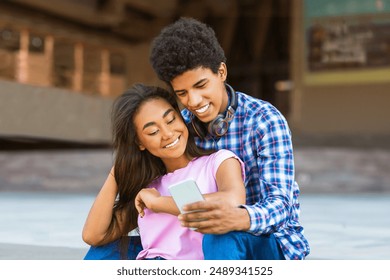 A teen black couple sits together on a concrete surface in an urban environment. Guy is wearing a blue and white plaid shirt and jeans, while the girl wears a pink T-shirt. They both appear happy - Powered by Shutterstock