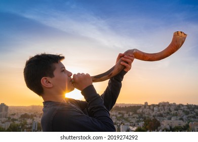 Teen boy blowing Shofar - ram's horn traditionally used for Jewish religious purposes, including the Feast of trumpets, Yom Kippur and Rosh Hashanah; beautiful sunset sky with sunburst in bachground