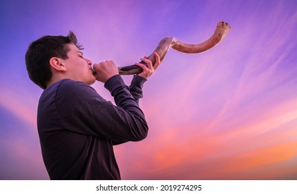 Teen boy blowing Shofar - ram's horn traditionally used for Jewish religious purposes, including the Feast of trumpets, Yom Kippur and Rosh Hashanah; beautiful pink sunset sky in bachground