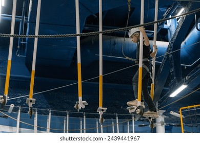 Teen boy carefully navigates an elevated rope obstacle wearing safety gear against an indoor backdrop - Powered by Shutterstock