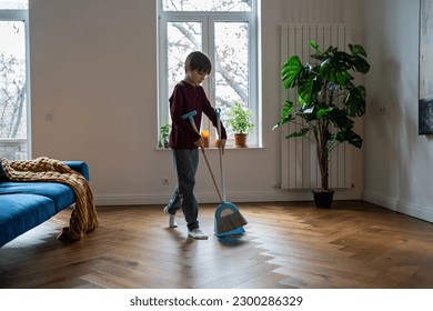 Teen boy doing chores cleaning floor in living room sweeping trash with broom to scoop. Teenager helping with household duties. Tidying up house involving children in family to maintain order at home. - Shutterstock ID 2300286329
