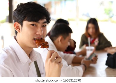 Teen Boy Eating Grilled Pork Or Barbecue With Friends While Having Socializing Activity In The University Campus, Selective Focused Picture Of A Lad Taking A Bite On Tasty Meat, Lifestyle Concept Pic