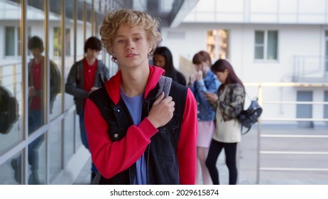 Teen Boy Looking At Camera With Classmates Talking On Background. Portrait Of Smiling Caucasian Schoolboy Posing At Camera Outside School