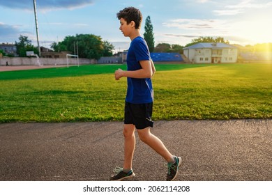 Teen Boy Running Along The Stadium Track, A Soccer Field With Green Grass - Concept Of Sports And Health