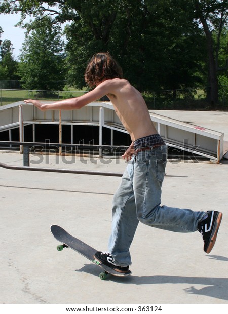 Teen boy shirtless in jeans skateboarding at outdoor skatepark. 