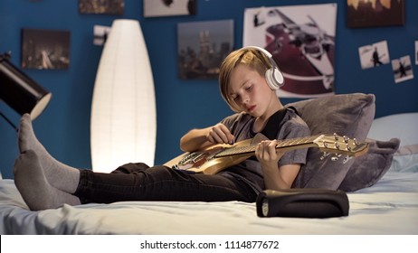 Teen boy sitting on bed at home and listening to music in headphones while practicing guitar