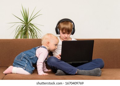 Teen Boy Sitting On Sofa With Laptop And Baby Girl Looks Curiously At The Monitor. Generation Alpha Concept.