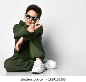 Teen boy in sportswear and sunglasses sitting on a gray background. Boy in a green suit shows a peace sign sitting near a free space for text. Concept of stylish sportswear for teenage children. - Powered by Shutterstock