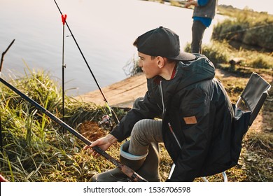 Teen Caucasian Boy Sit Near Lake On Fishing Stool And Try Fish.