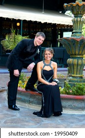 Teen Couple Sit Outdoors At A Water Fountain Dressed And Ready For Prom Night.  Tux And Gown Both In Black.