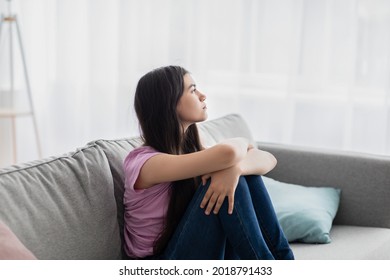 Teen Depression Concept. Stressed Indian Teenage Girl Sitting On Couch At Home, Looking Out Window, Suffering From Lack Of Communication, Feeling Upset And Lonely During Covid Lockdown