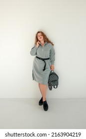 Teen Fashion Leisure. Young Plus Size Redhead Girl Is Standing Casual In Gray Oversize Dress And Smiling Cute With Backpack In Hand On The White Wall Background. Fashion Concept, Free Space