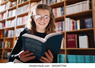 Teen girl among a pile of books. A young girl wearing glasses reads a book with shelves in the background. She is surrounded by stacks of books. Book day. - Powered by Shutterstock